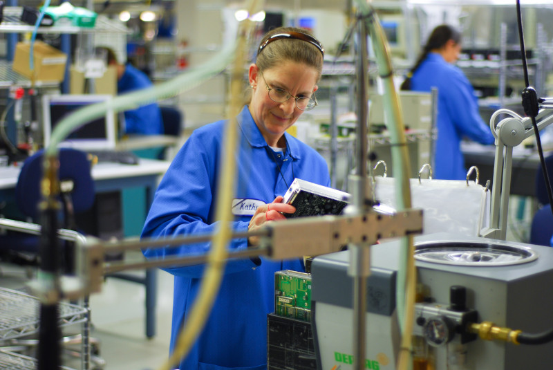 Factory worker in a blue uniform examines a circuit board in an electronics manufacturing setting. The workspace is filled with various equipment and machinery.