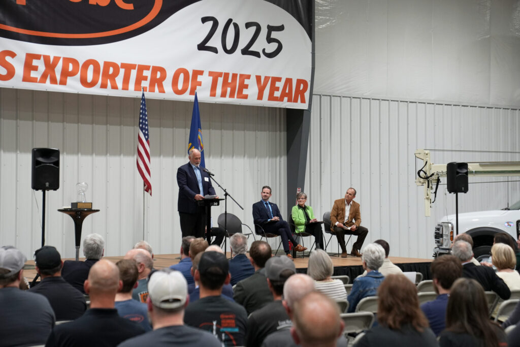 A speaker addresses an audience at a podium under a large "2025 Exporter of the Year" banner. Two flags are behind the podium. Three individuals are seated on the stage. The event takes place in a large indoor space with a vehicle partially visible on the right.