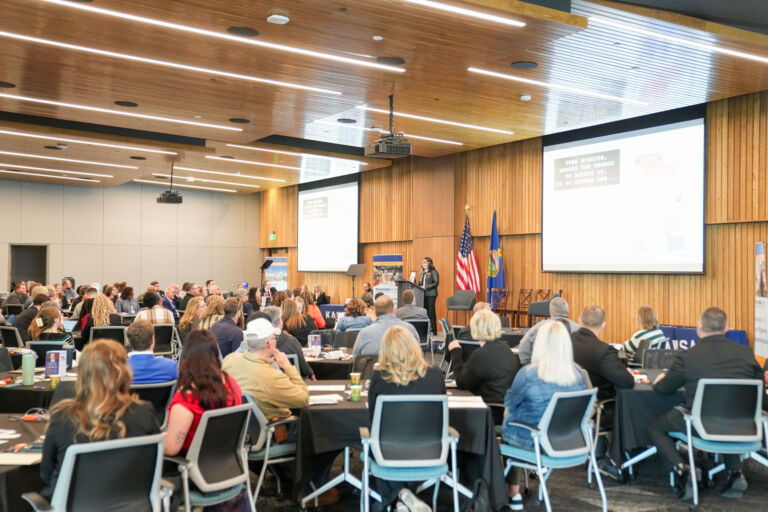 A large conference room with many attendees seated at tables, facing a presentation screen. Two flags are visible: the United States flag and another flag. The room features wooden paneling and high ceilings.