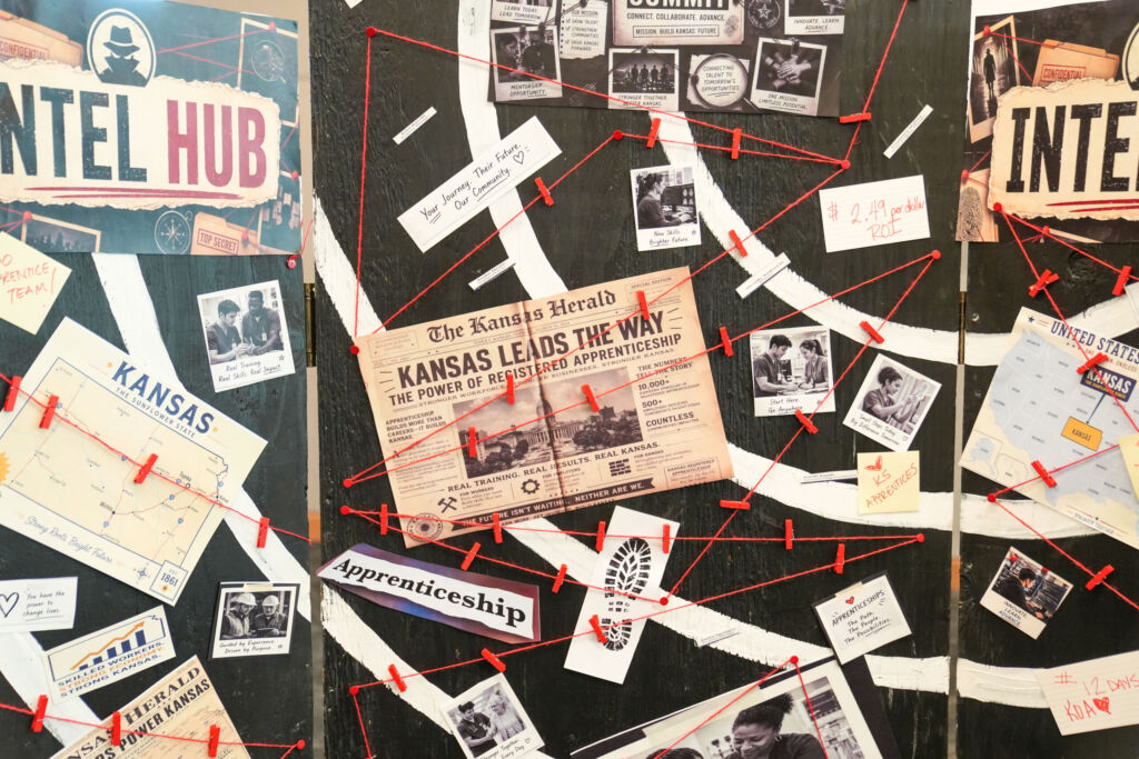 A bulletin board is filled with various newspaper clippings, photographs, and documents connected by red string. Prominent elements include a newspaper headline from "The Kansas Herald" reading "Kansas Leads The Way" about registered apprenticeship, and the logos for the "United States Department of Labor" and "Intel Hub." The board appears to depict a complex, interconnected investigation or project.