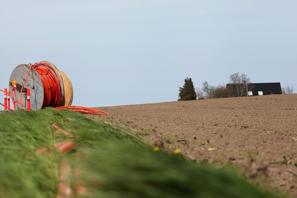A large spool of orange cable is positioned on the edge of a cultivated field. A small black-roofed building is visible in the distance. The sky is clear.