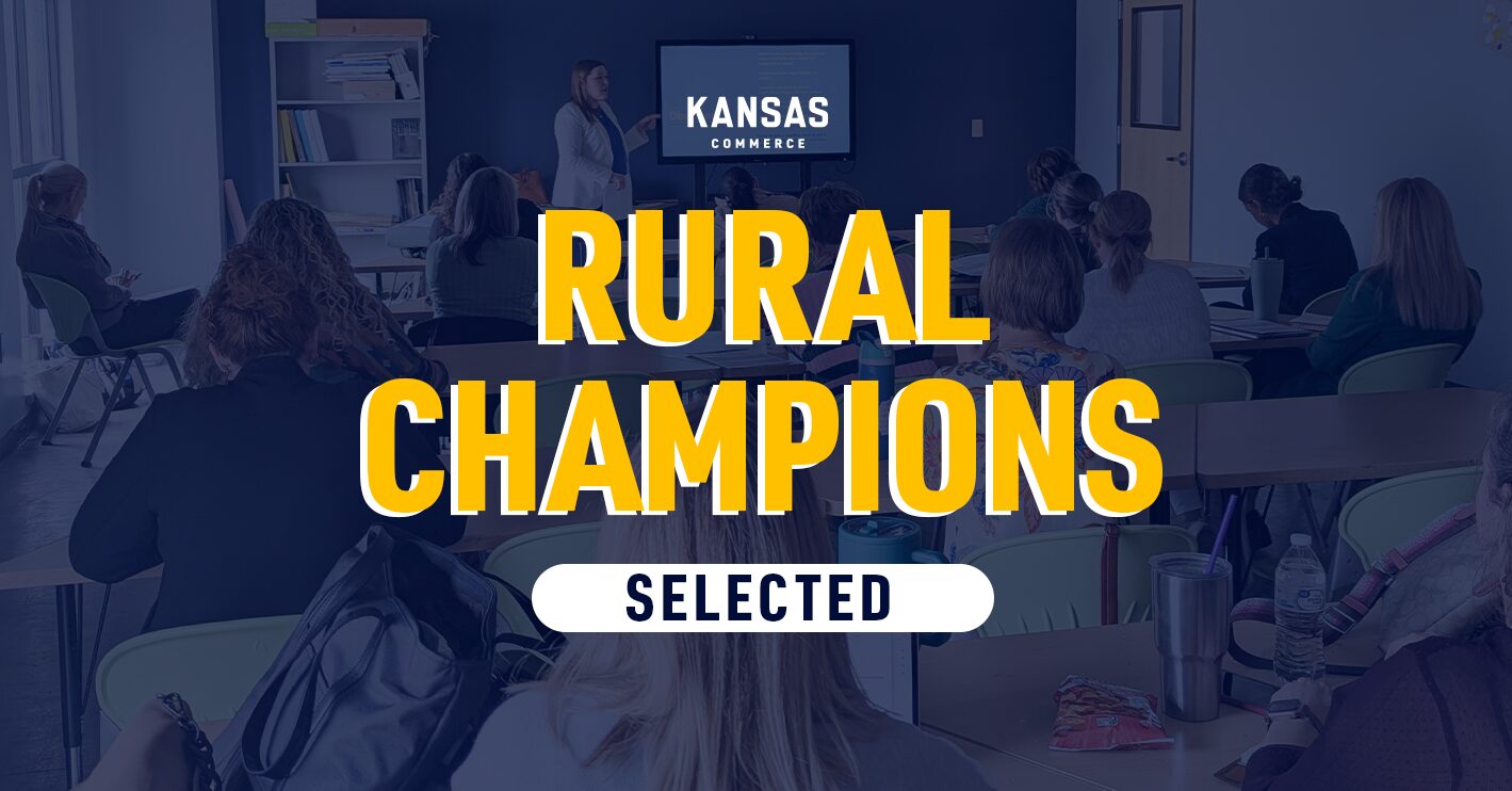 A group of people sitting in a classroom setting, facing a screen displaying "Kansas Commerce." The text "Rural Champions Selected" is prominently displayed over the image.