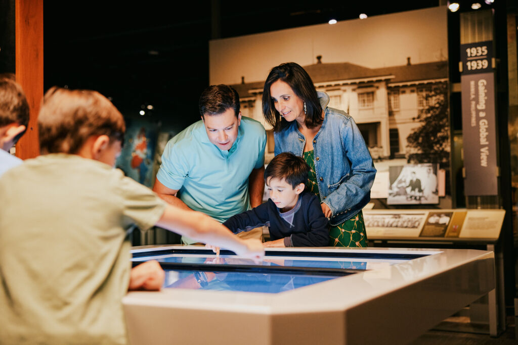A group of people engage with an interactive display at a museum. Historical photos and exhibits are visible in the background.