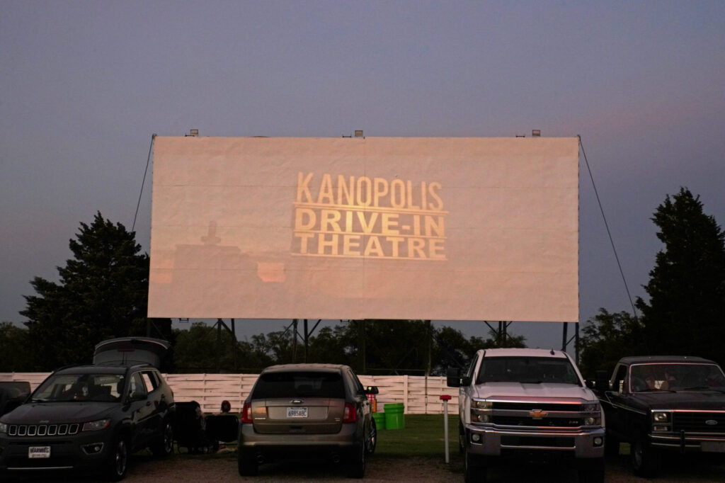 A drive-in theater screen displays the words "Kanopolis Drive-In Theatre" at dusk. Several cars are parked in front of the screen, surrounded by trees and a clear sky.
