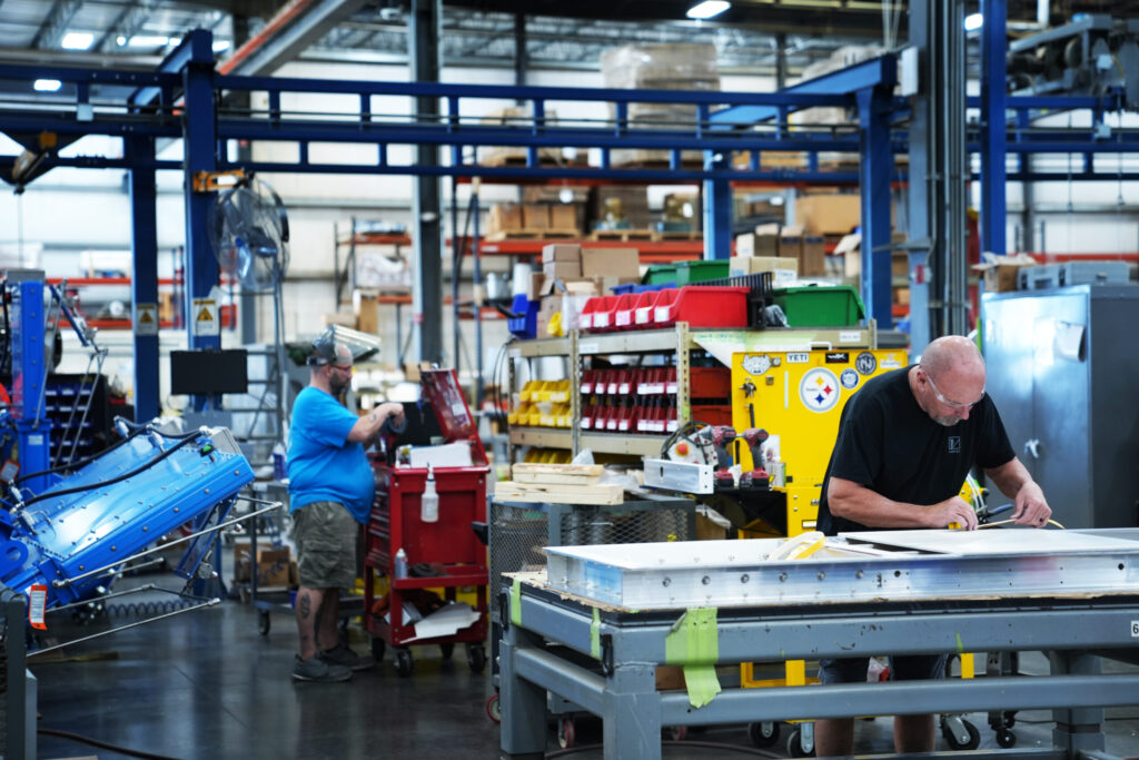 A busy workshop with two individuals working. One person is handling a tool cart, and another is working at a table. Shelves filled with various tools and equipment are visible in the background. The setting has bright lighting and industrial decor.
