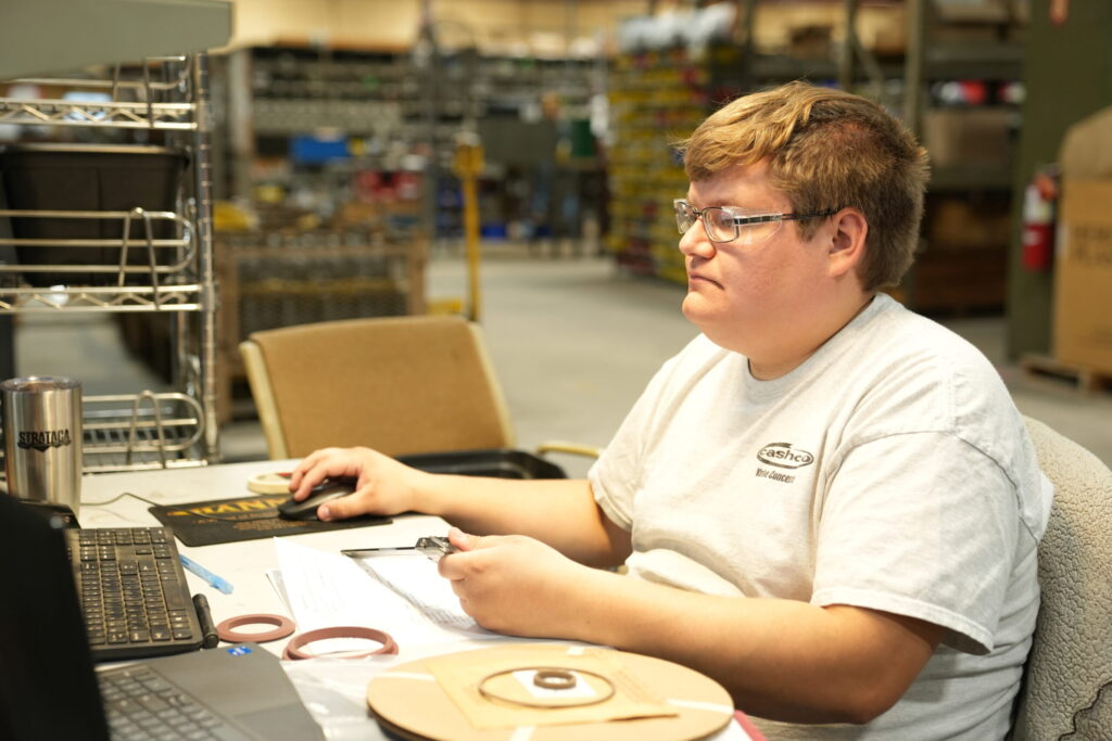 An individual at a workstation in a warehouse setting, wearing glasses and a white "Swagelok" branded shirt, is working on a computer. The desk is cluttered with papers, a tape dispenser, and a metallic tumbler. Shelving units and industrial supplies are visible in the background.