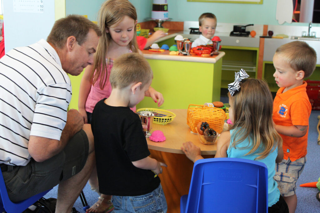 A group of people, including an adult and children, are gathered around a small table in a colorful playroom. The table has toy kitchen items. A child in the background is playing near a play kitchen setup.