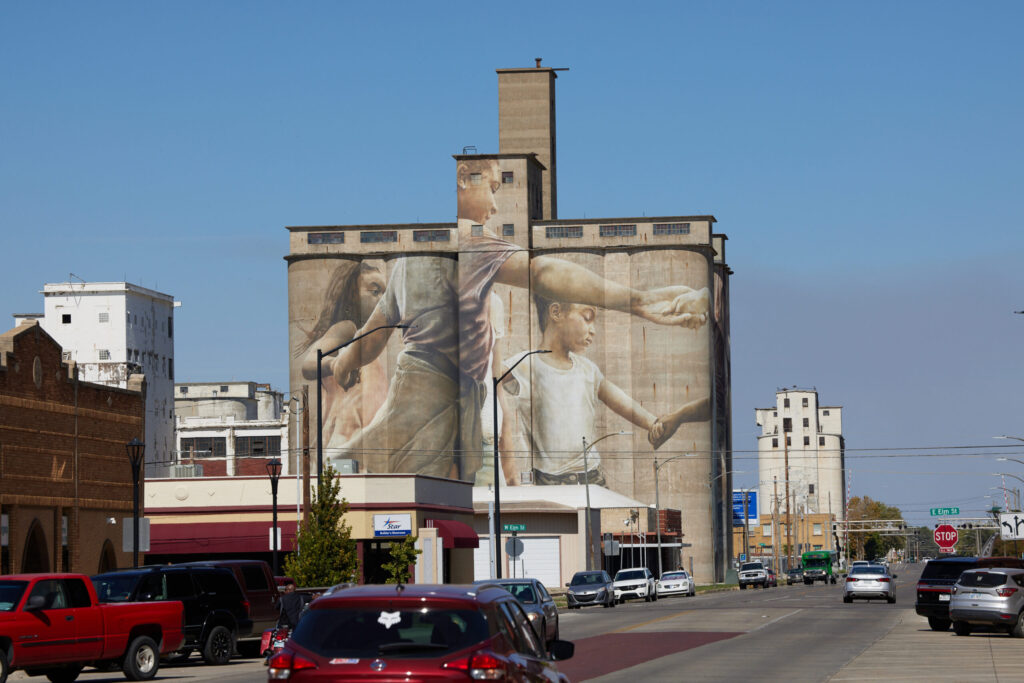A large mural covers the side of a tall concrete grain elevator in a town setting. The mural depicts two people, one of whom appears to be an adult and the other a child, reaching out to each other. There are several cars along the street, and buildings are visible in the background. The sky is clear and blue.