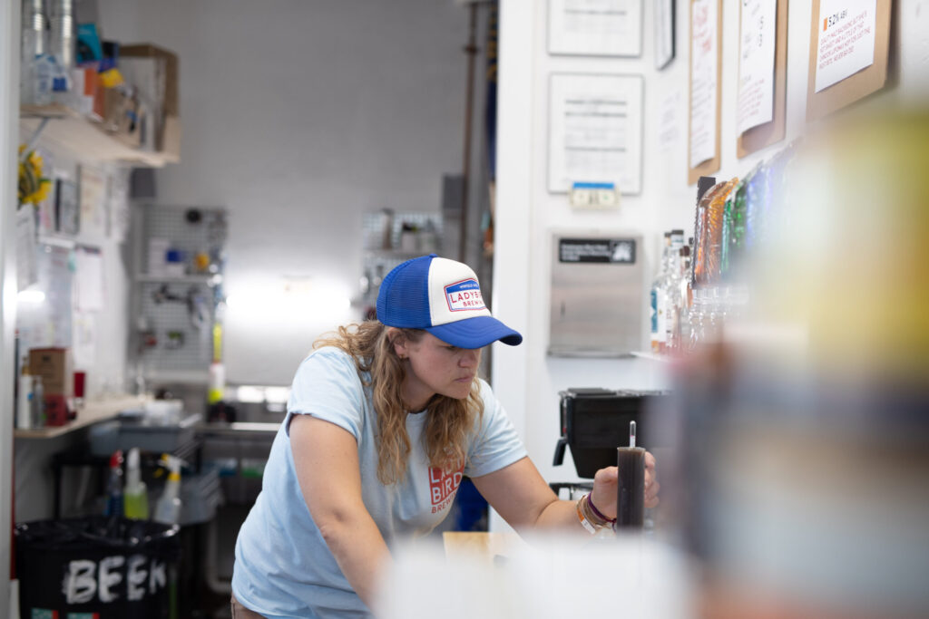 A person wearing a baseball cap labeled "Double Mountain Beer" is working at a counter in a brewery. The room has various equipment and a menu on the wall.