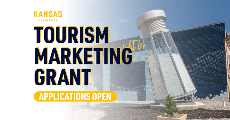 A tilted large metallic structure resembling a salt shaker stands in front of a blue mural under a clear sky. The text reads, "KANSAS COMMERCE TOURISM MARKETING GRANT APPLICATIONS OPEN." There are small plants near the base of the structure.