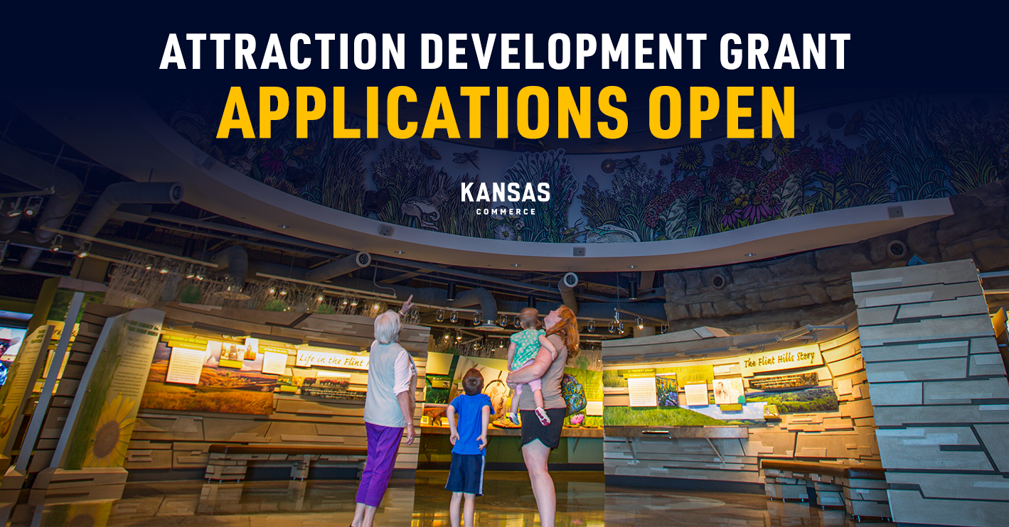 A group of people stand in a museum-like environment, looking around at displays. Above them, a banner reads "Attraction Development Grant Applications Open" with "Kansas Commerce" below it. The walls are adorned with various exhibits and information panels.