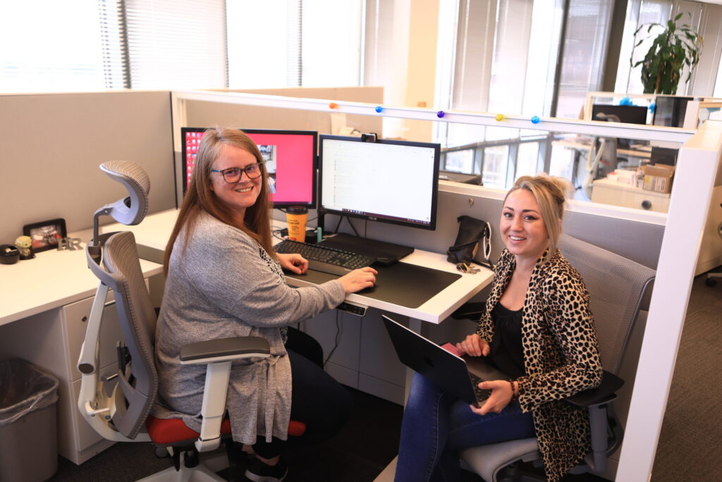 Two people are seated in an office cubicle, both facing the camera and smiling. One person is working at a desktop computer with a second display showing various applications, while the other holds a laptop. The cubicle is decorated with string lights, and there are large windows in the background letting in natural light.