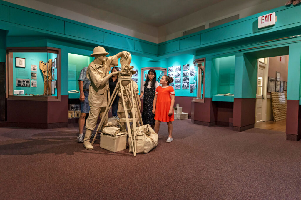 A group of people stand in a museum exhibit, observing a statue of an early filmmaker with a vintage camera. The display includes historical photographs and artifacts, with teal walls creating a colorful backdrop. The setting combines educational and artistic elements.