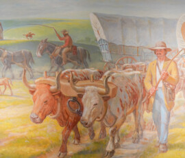 A painting depicts pioneers traveling with a covered wagon. Two oxen pull the wagon, guided by a person walking alongside. In the background, another person rides a horse, herding additional oxen. A dog trots nearby, and a small house is visible in the distance. The scene is set in a grassy, open landscape.