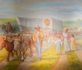 A pastoral scene illustrating a group of pioneers traveling west with covered wagons. Oxen are pulling the wagons across a sunlit prairie. Travelers, including adults and children, walk alongside. The landscape is lush and green, capturing a sense of movement and hope.