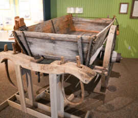 A wooden wagon displayed in a museum setting, mounted on a stand. The wagon appears aged, with weathered wood and iron fittings. The background includes framed documents and informational displays.