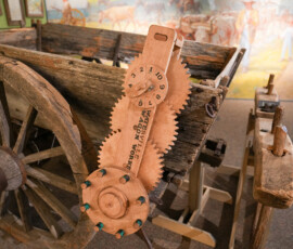 A wooden wagon featuring detailed mechanical workings is displayed in a museum setting. A wooden gear system is attached to the wagon's side, labeled with "Watson's Wagon Works Minneapolis." The background shows a mural depicting historical scenes with figures and horses.