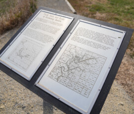 A display stand with two informational plaques. The left plaque features a map titled "Public Roads and Bridges in the Middle District," with text above it. The right plaque includes historical text and a map marked "Davis County." The stand is set on a gravel path with grass visible in the background.