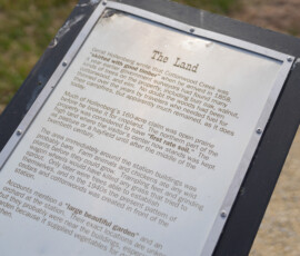 A close-up of an information board describing the history of the land. The text mentions Garrett Hollenbeck's 160-acre claim in 1855, the development of the area, and the types of trees present, such as walnut, hickory, and oak. It also details the presence of a large garden and the use of lumber for construction. The sign is mounted on a stand next to a gravel path.