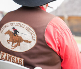 A person wearing a brown vest with a "National Pony Express Association" patch and the word "Kansas" sits on a white chair. The vest features an embroidered image of a rider on a galloping horse. A blurred building is in the background.