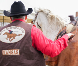 A person wearing a cowboy hat and a vest with a "National Pony Express Association" logo is standing beside a horse. The vest has the word "Kansas" on it. There is a tent in the background.