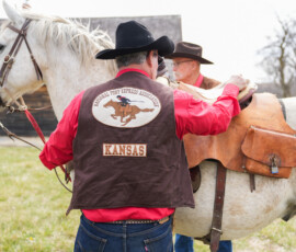 Two individuals are outdoors with a white horse. One is adjusting the saddle. They are wearing cowboy hats and attire featuring "National Pony Express Association" and "Kansas" on the vest. A wooden building is in the background.