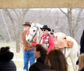 Pony Express riders stand next to horse and listen to speaker at Pony Express Byway dedication event.