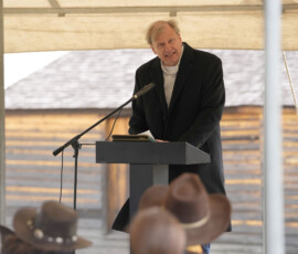 Speaker stands at a podium under a tent addressing an audience at a Pony Express event