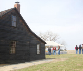 Individuals stand outside outside an old cabin.